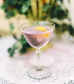 In a clear cocktail up glass, a light purple cocktail with a lemon peel garnish is featured.