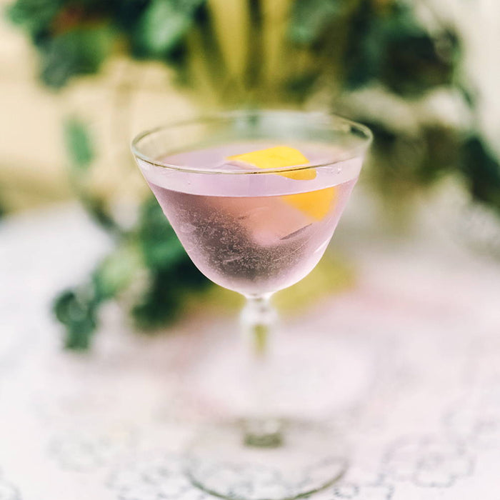 In a clear cocktail up glass, a light purple cocktail with a lemon peel garnish is featured.