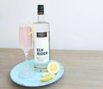 A small light blue plate holds a bottle of HDC Elk Rider Gin, a light pink cocktail in a flute, and some lemon wedges.