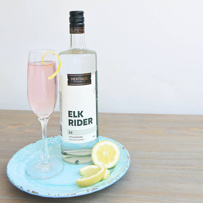 A small light blue plate holds a bottle of HDC Elk Rider Gin, a light pink cocktail in a flute, and some lemon wedges.