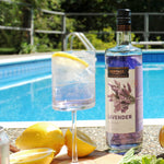 A bottle of HDC Lavender Vodka is poolside with the Sparkling Lavender Cocktail in a glass of ice next to it. There is also a cut lemon in the photo with a lemon slice in the drink itself.
