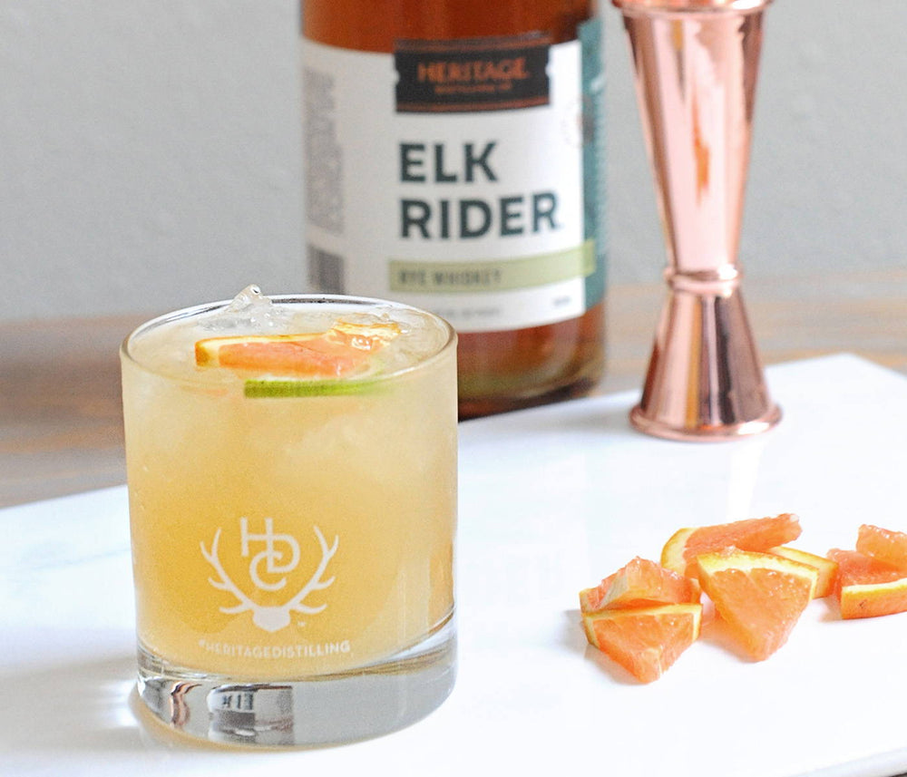 A bottle of HDC Elk Rider Rye Whiskey, a copper jigger, and slices of grapefruit are displayed. In front, is a yellow colored cocktail in a Heritage Distilling Co. tumbler of ice with grapefruit and lime as garnishes 