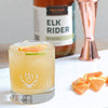 A bottle of HDC Elk Rider Rye Whiskey, a copper jigger, and slices of grapefruit are displayed. In front, is a yellow colored cocktail in a Heritage Distilling Co. tumbler of ice with grapefruit and lime as garnishes 