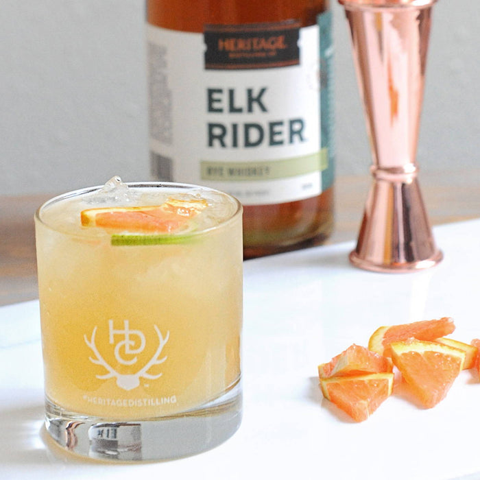 A bottle of HDC Elk Rider Rye Whiskey, a copper jigger, and slices of grapefruit are displayed. In front, is a yellow colored cocktail in a Heritage Distilling Co. tumbler of ice with grapefruit and lime as garnishes 