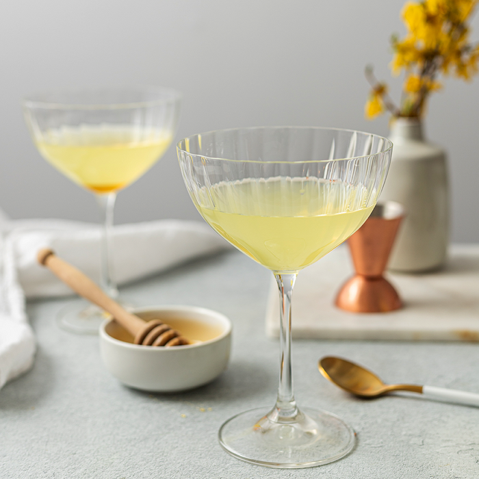 Two cocktail glasses filled halfway up to the top with a light yellow cocktail and honey simple syrup displayed in a dish in the background.
