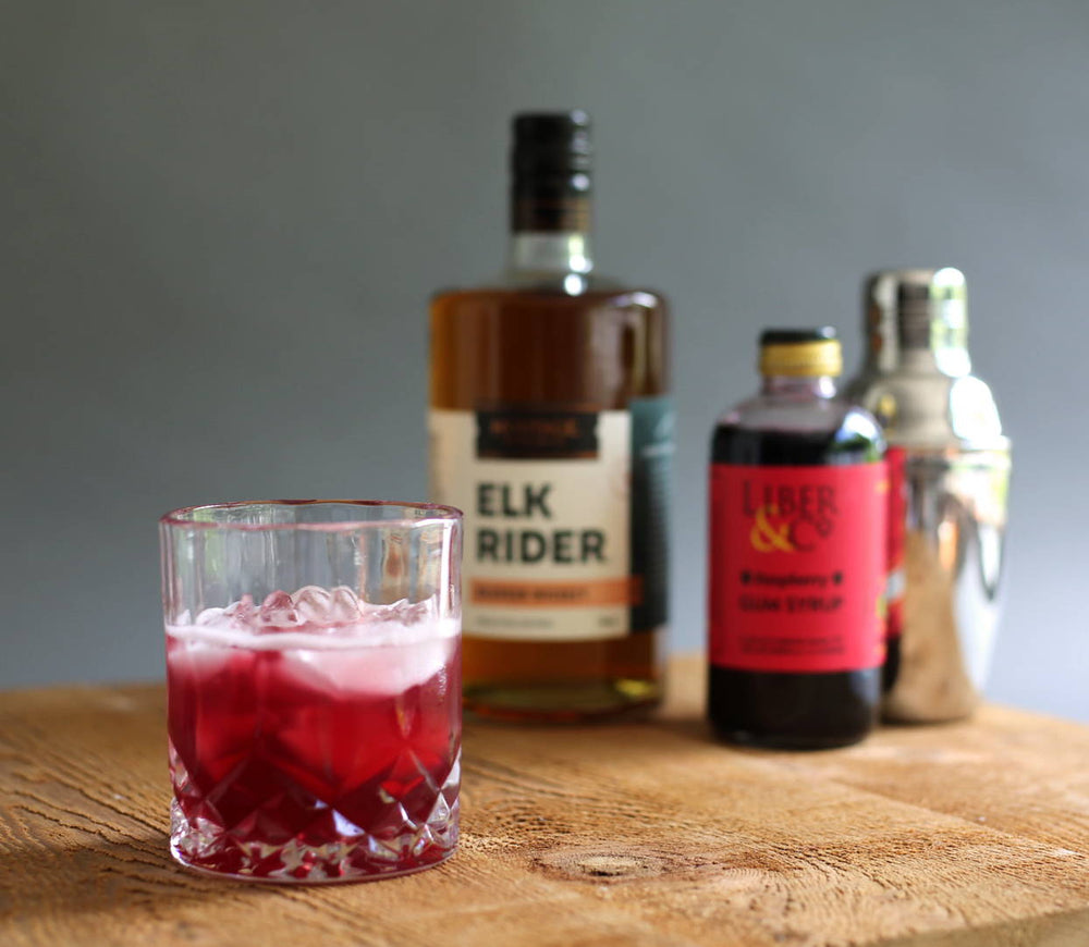 A deep red colored cocktail in a tumbler glass of ice. In the background HDC Elk Rider Bourbon, Liber & Co. Raspberry Gum Syrup, and a metal shaker are featured as well.