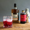 A deep red colored cocktail in a tumbler glass of ice. In the background HDC Elk Rider Bourbon, Liber & Co. Raspberry Gum Syrup, and a metal shaker are featured as well.