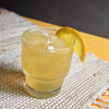 A yellow rum-based cocktail (using HDC Commander's Spiced Rum) in a glass of ice with a lemon wedge garnish and photographed on top of a woven table runner.