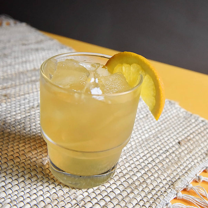 A yellow rum-based cocktail (using HDC Commander's Spiced Rum) in a glass of ice with a lemon wedge garnish and photographed on top of a woven table runner.