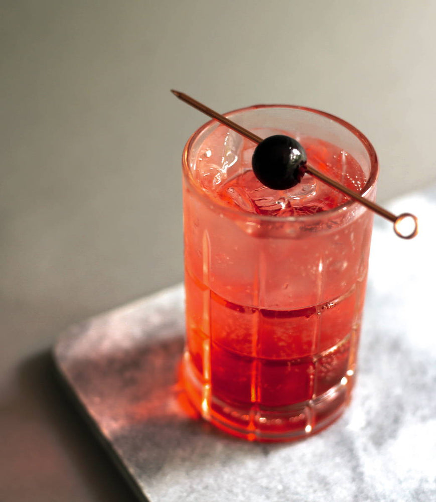 A tall tumbler of ice with a pinkish red cocktail and a Luxardo cherry garnish on a stir stick.