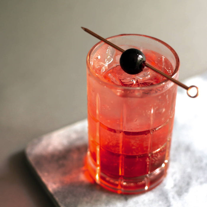 A tall tumbler of ice with a pinkish red cocktail and a Luxardo cherry garnish on a stir stick.