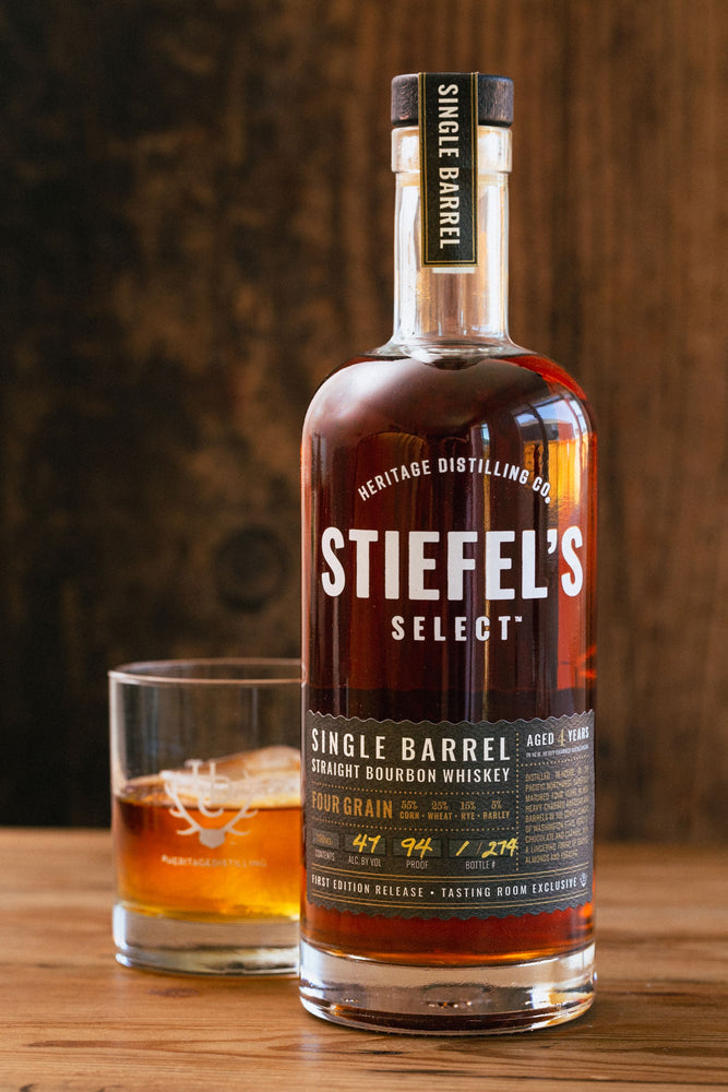 
                  
                    A 750ml glass bottle of whiskey "Stiefel's Select Single Barrel Four Grain Bourbon" sitting atop a wooden counter next to a short rocks glass filled with whiskey. 
                  
                